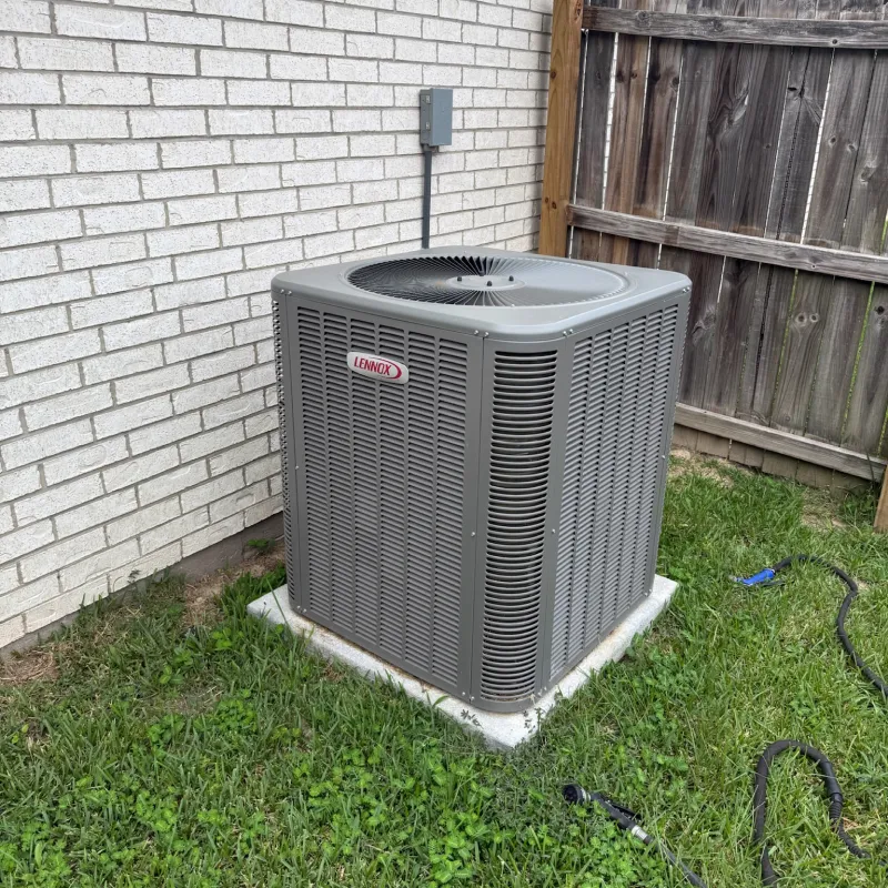A gray Lennox HVAC unit on a concrete slab next to a white brick wall and wooden fence, with green grass.
